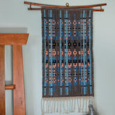 The Northern Colors Bound Weave tapestry by Halcyon Yarn, featuring a bound weave pattern in blue, peach, and black tones, is displayed against a light wall. The woven wall hanging is suspended from a horizontal wooden rod, and fringe adorns the bottom edge. A wooden structure is partially visible on the left side.