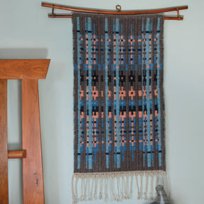 The Northern Colors Bound Weave from Halcyon Yarn is a woven wall hanging that features a bound weave pattern in shades of blue, orange, and black. It is mounted on a wooden rod and hangs against a light-colored wall, with a partially visible wooden structure on the left. Cream-colored fringe adorns the bottom of this elegant textile.