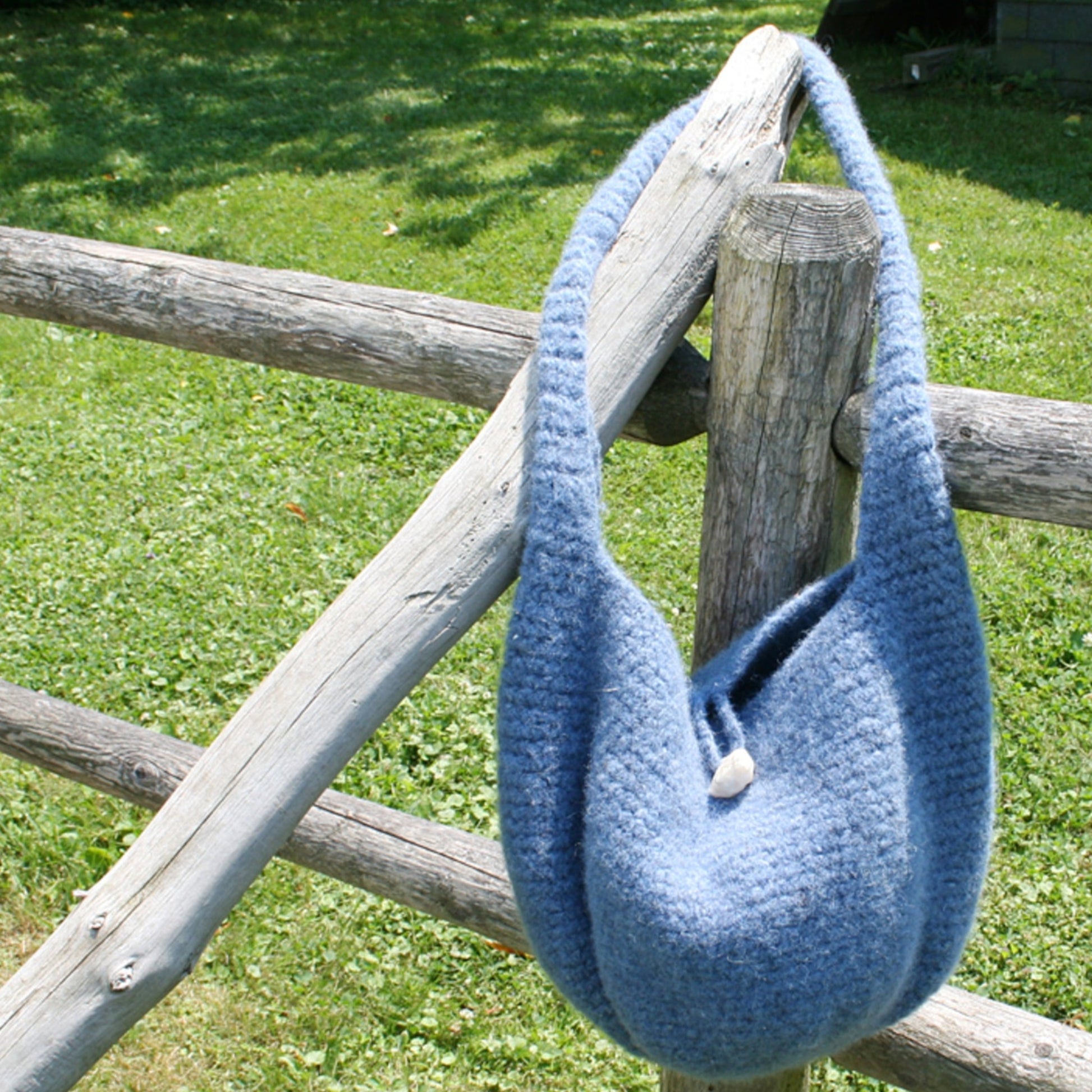 The Crochet Felted Satchel, a handmade bag crafted with Halcyon Yarn, hangs on a rustic wooden fence post in a grassy outdoor area on a sunny day. The textured blue woolen bag features a single button closure.