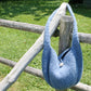 The Crochet Felted Satchel by Halcyon Yarn, featuring a stone button closure, hangs on a rustic wooden fence in a grassy area, with the scene bathed in sunlight.