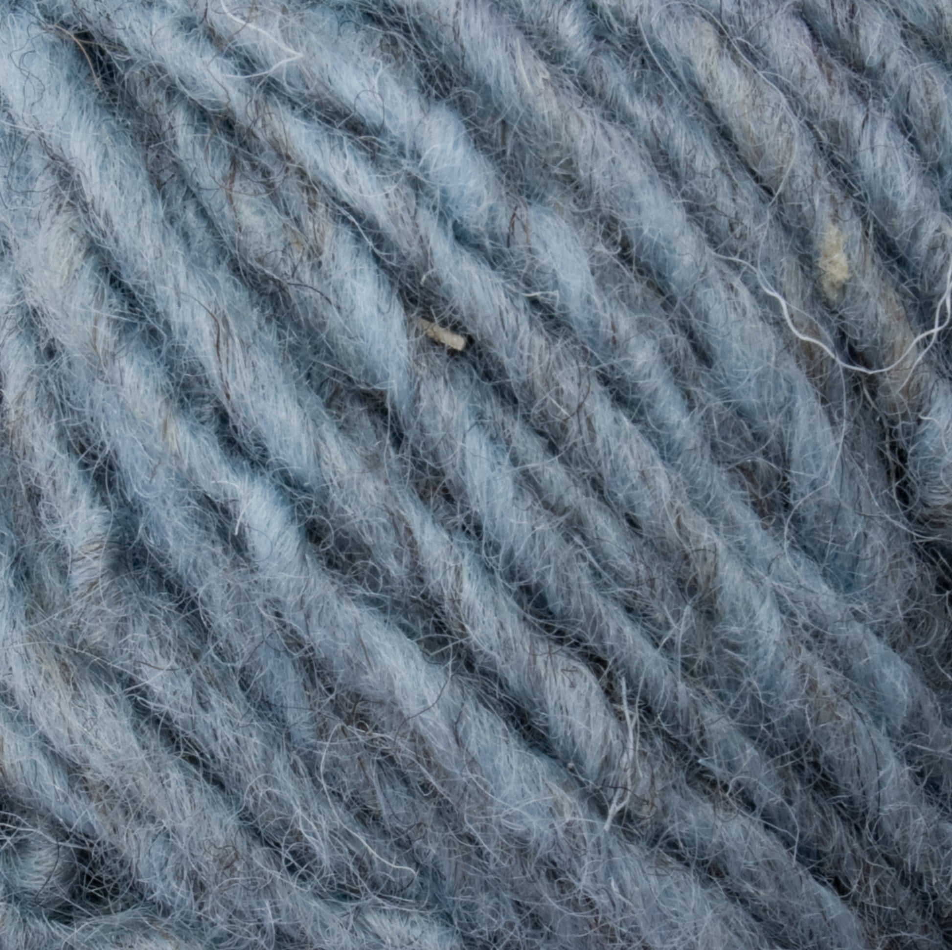 Close-up view of a textured gray and blue Halcyon Geo Rug Wool from Caledonian Dye Works, featuring tightly coiled fibers. The image showcases the intricate details of the strands, illustrating the wool's soft and fuzzy appearance.