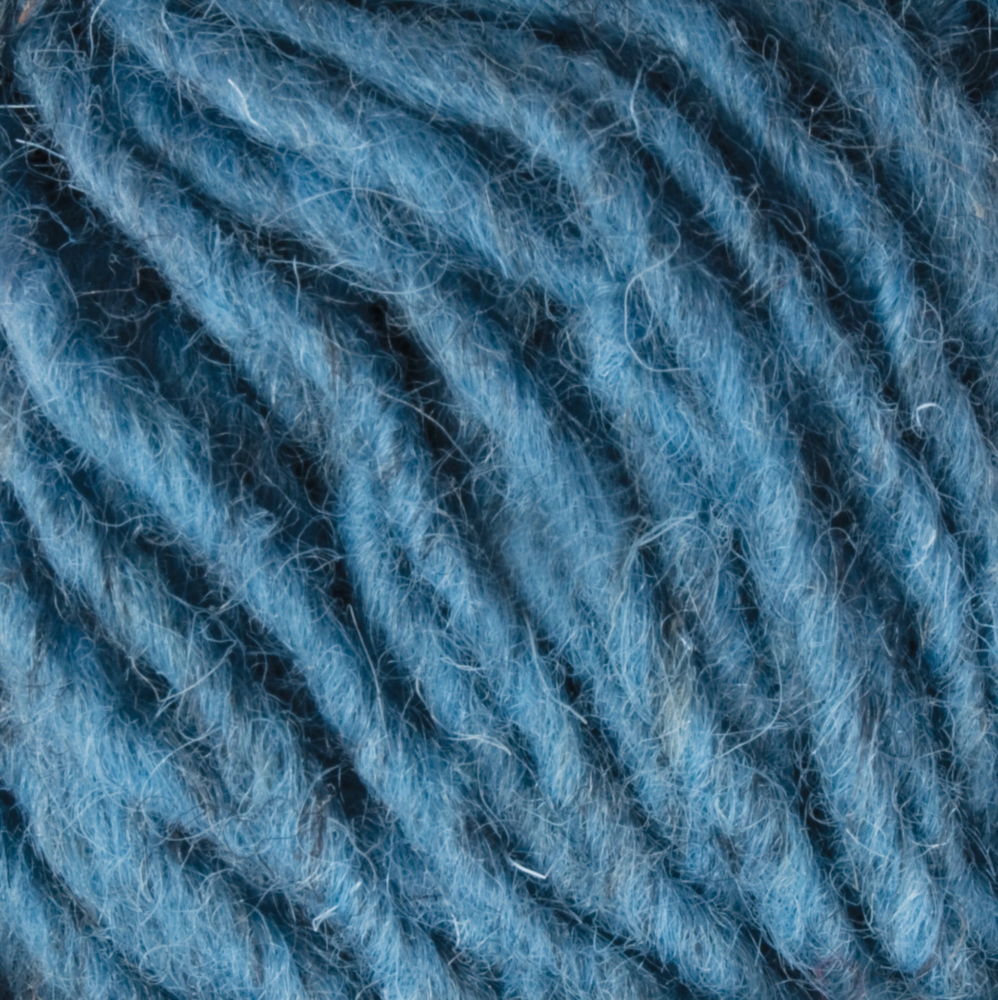 Close-up image of soft, blue Halcyon Geo Rug Wool fibers intertwined and textured. The strands appear thick and fuzzy, with varied shades of blue throughout. The texture and detail of the fibers are prominent, showcasing the intricate pattern of the yarn by Caledonian Dye Works.