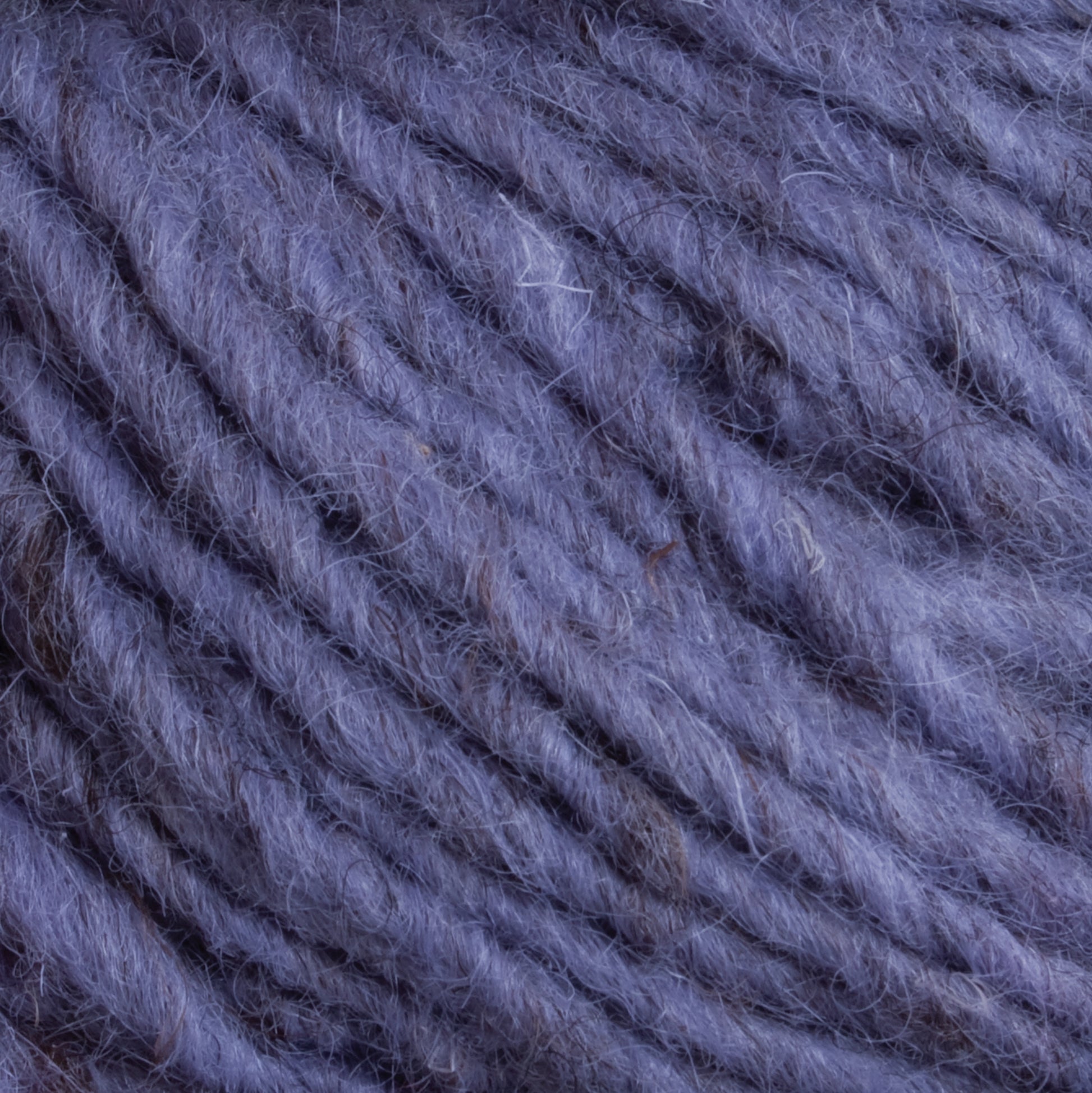 Close-up image of soft, chunky, purple yarn with visible individual strands and fibers entwined together. The texture appears fuzzy and thick, reminiscent of Caledonian Dye Works' Halcyon Geo Rug Wool, making it suitable for knitting or crocheting cozy items.