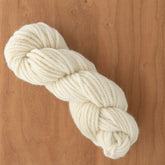 A skein of Lemieux's Halcyon Yarn 4-ply Rug Wool, naturally dyed to enhance its cream-colored hue, is neatly twisted into a coil and placed on a wooden surface. The yarn appears soft and thick, suitable for knitting, crocheting, or even latch hooking projects.