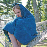 A person sits on a hammock in a wooded area, wrapped in the Casco Cottage Knitted Throw from Halcyon Yarn, complete with a cozy hood, looking down pensively. The surrounding trees and greenery create the perfect backdrop for this versatile blanket.