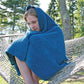 A person with light hair sits on a large hammock outdoors, wrapped tightly in a Casco Cottage Knitted Throw from Halcyon Yarn, complete with a hood. The surrounding area has trees and green grass, suggesting a peaceful but cool environment. The person appears thoughtful or contemplative.