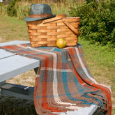 A heathered Harrisville picnic basket sits on a gray picnic table with a green hat on top. A cozy Woven Fall Picnic Blanket from Halcyon Yarn, featuring plaid shades of blue, orange, brown, and white, is draped over the table. Nearby sits a green apple. The background includes grass and greenery.