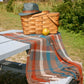 A woven picnic basket with a green hat on top sits on a blue wooden picnic table. The Halcyon Yarn Woven Fall Picnic Blanket, download, drapes over the table, showcasing its intricate weaving pattern. A yellow-green apple rests beside the basket, and lush greenery surrounds the peaceful outdoor setting.