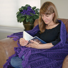 A woman with long brown hair is sitting in an armchair, wrapped in a Halcyon Yarn Undulating Waves Woven Blanket, reading a book. Behind her, there is a potted plant with purple flowers on a table. The room has neutral-colored walls and a window with blinds.