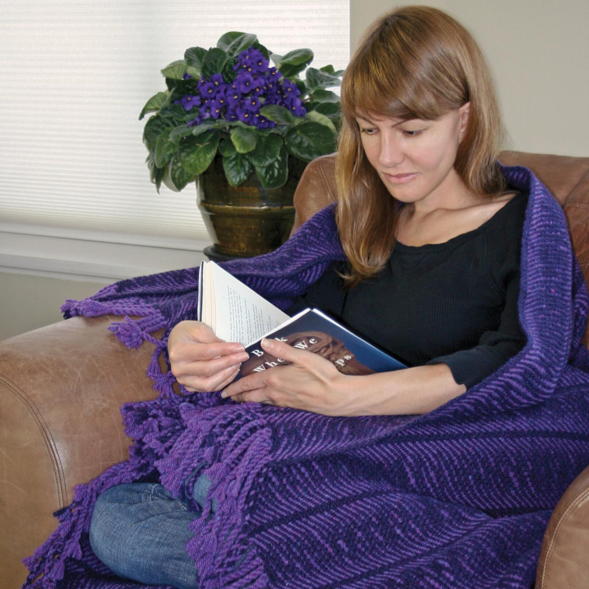 A woman with long brown hair sits in a brown armchair, wrapped in the Halcyon Yarn Undulating Waves Woven Blanket, reading a book. Behind her, there is a potted plant with purple flowers on a table next to a window with closed blinds.