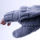 A close-up shot displays a hand adorned with Halcyon Yarn's Soldier's Glomitt. These gray knitted mittens are fingerless, allowing the thumb and tips of the fingers to peek through for easy texting. They also include a foldover mitten flap for additional warmth. The background features a soft, neutral color.