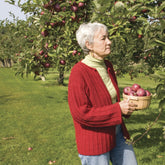 An elderly person with short white hair, dressed in a Graceful Ribbed Cardigan by Halcyon Yarn over a light green turtleneck sweater, stands in an orchard holding a basket of red apples. They are looking intently at the apples growing on the tree beside them. The background reveals lush green grass and more apple trees, creating an idyllic scene that would inspire any knitter.