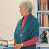 An elderly woman with short gray hair is pictured in profile, wearing a red top paired with Halcyon Yarn's Asymmetrical One-Button Wrap. She stands next to a bookshelf filled with books and several decorative items, including a round sculpture and a small statuette.