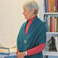 An elderly woman with short gray hair is pictured in profile, wearing a red top paired with Halcyon Yarn's Asymmetrical One-Button Wrap. She stands next to a bookshelf filled with books and several decorative items, including a round sculpture and a small statuette.