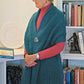 An elderly woman with short gray hair stands in profile in front of a bookshelf. She is wearing a teal Asymmetrical One-Button Wrap by Halcyon Yarn over a red turtleneck sweater, clasped with an off-set button closure. The bookshelf is filled with books and various decorative items.