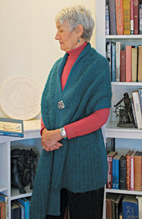 An elderly woman with short gray hair stands in profile in front of a bookshelf. She is wearing a teal Asymmetrical One-Button Wrap by Halcyon Yarn over a red turtleneck sweater, clasped with an off-set button closure. The bookshelf is filled with books and various decorative items.