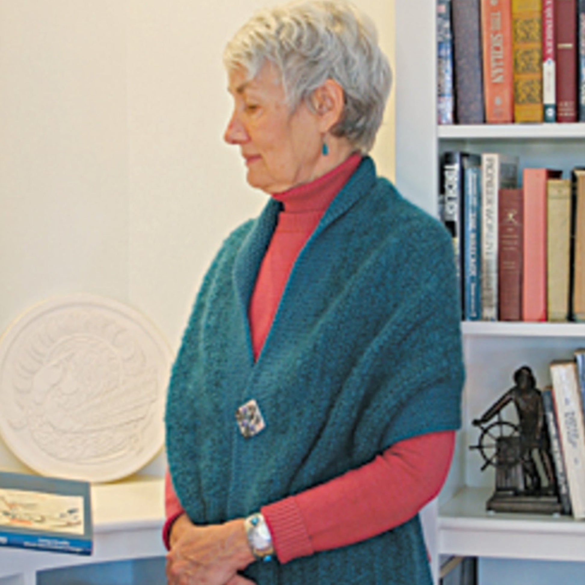 An elderly woman with short gray hair is standing and gazing sideways, clad in a red turtleneck and the Halcyon Yarn Asymmetrical One-Button Wrap, secured with a brooch. She is in a room featuring white walls and a bookshelf in the background, filled with an array of books and decorative items.