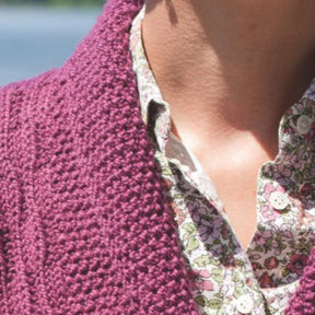 Close-up of a person wearing the Wellfleet Jacket by Twist Collective in a rich purple hue, layered over a white shirt with a floral pattern. The image is cropped to show only part of the neck, shoulders, and upper chest. The background is slightly blurred and suggests an outdoor setting.