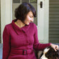 Wearing a Mod Podge Jacket Pattern by Halcyon Yarn, a person sits on a porch, gently petting a brown and white dog. With a slight smile, they gaze down at the dog. Behind them is a closed door and walls with green paneling.