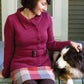 A woman with short dark hair sits on a porch, petting a black, brown, and white dog. She is wearing the Mod Podge Jacket by Halcyon Yarn – a red knit cardigan with seed stitch borders and subtle waist shaping. Accompanying her ensemble are a plaid skirt and dark tights. The background features a partially visible door and part of a house façade.