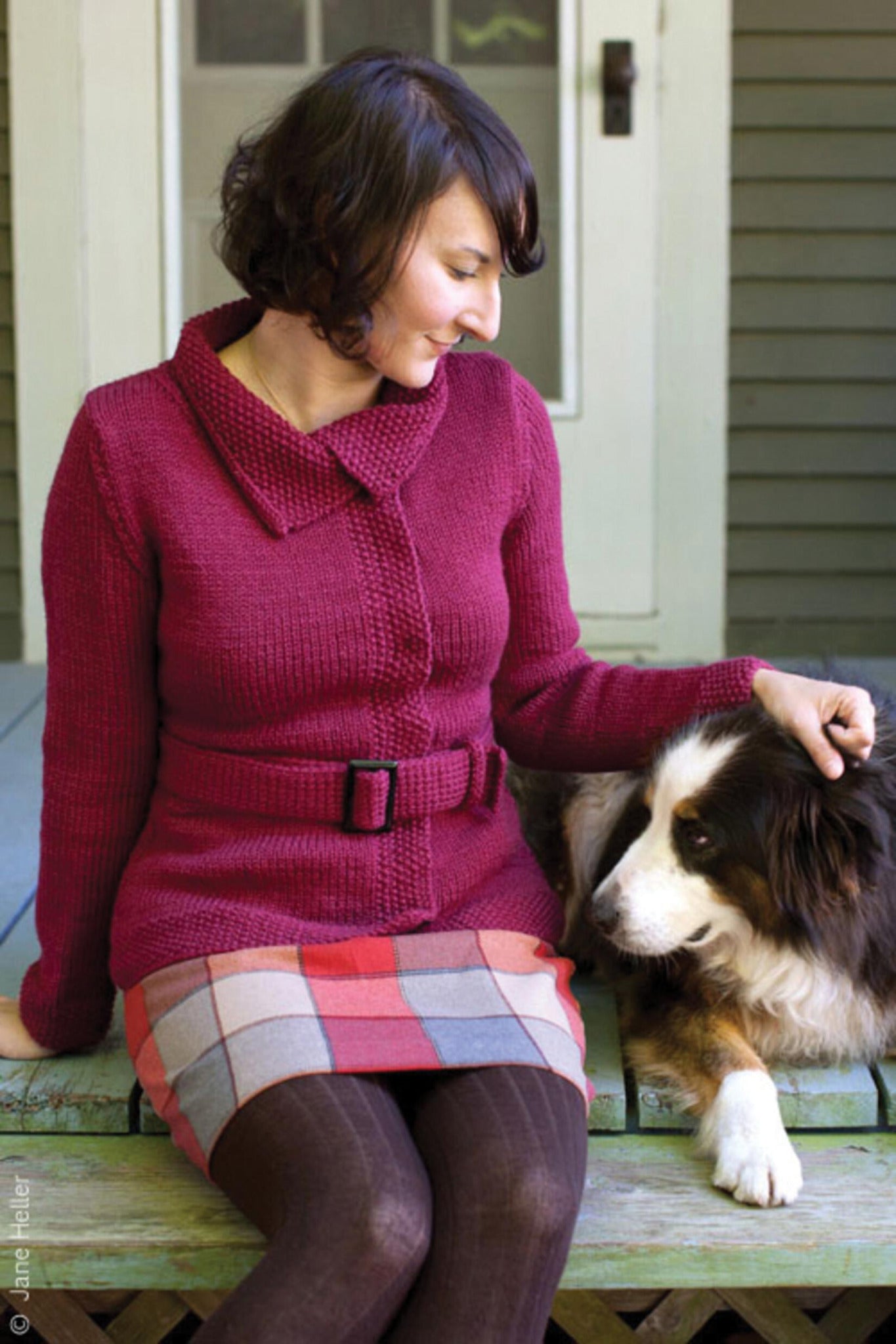 A woman with short dark hair sits on a porch, petting a black, brown, and white dog. She is wearing the Mod Podge Jacket by Halcyon Yarn – a red knit cardigan with seed stitch borders and subtle waist shaping. Accompanying her ensemble are a plaid skirt and dark tights. The background features a partially visible door and part of a house façade.