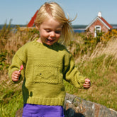 A smiling blonde-haired child, dressed in a green "Ship Shape Kid's Crew" pullover from Halcyon Yarn and a purple skirt, holds a small item in their hand while standing outside. In the background, tall grass, a red-roofed house, and a body of water under a clear blue sky are visible.