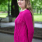 A woman with long hair, wearing a vibrant pink knitted sweater from Halcyon Yarn's Printed Pattern Roxton Pullover collection, smiles while standing outdoors in a park. Green trees and blurred background elements enhance the serene, natural setting.