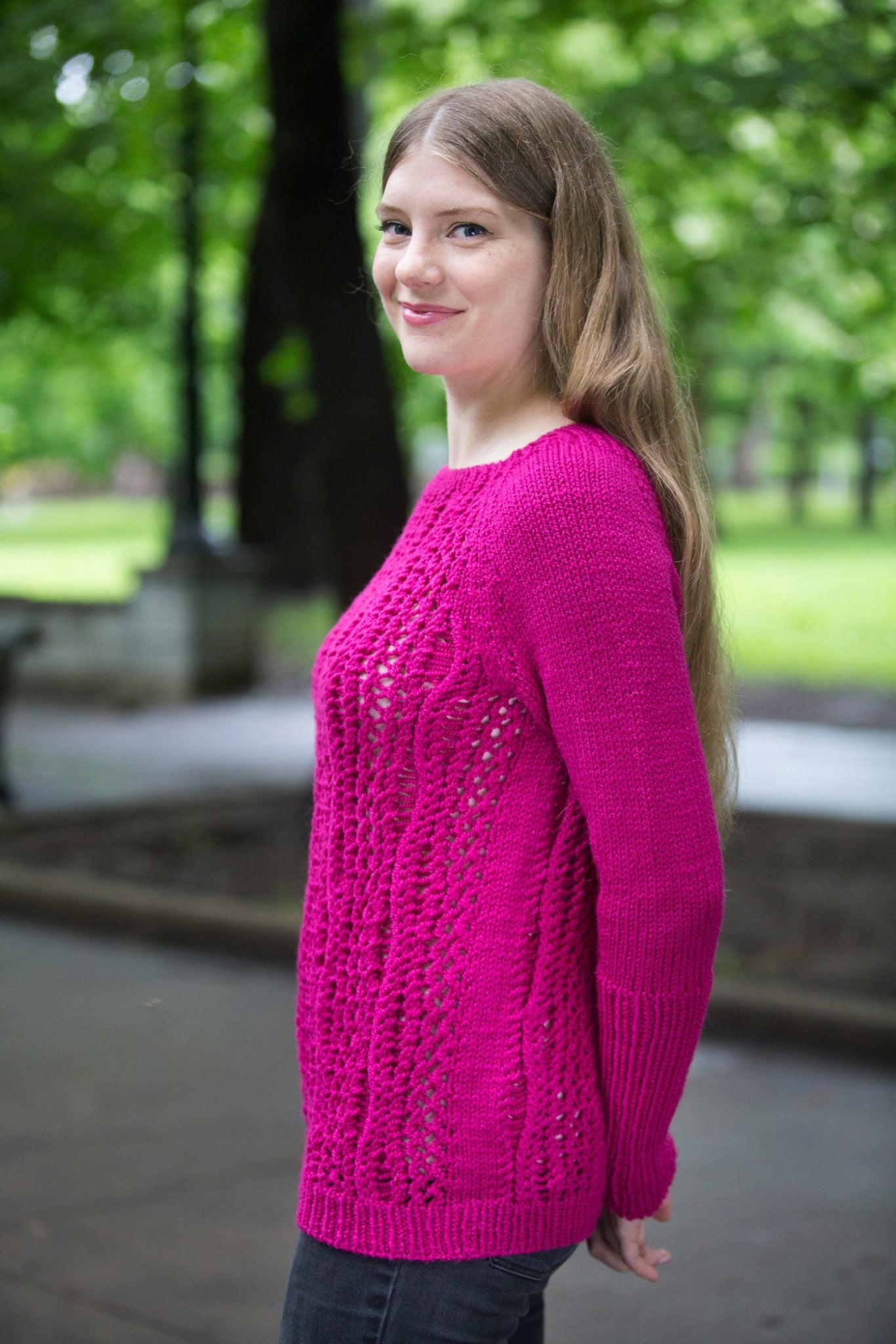 A woman with long hair, wearing a vibrant pink knitted sweater from Halcyon Yarn's Printed Pattern Roxton Pullover collection, smiles while standing outdoors in a park. Green trees and blurred background elements enhance the serene, natural setting.