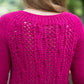 A person with long blond hair is seen from the back wearing a bright pink knitted sweater, much like Halcyon Yarn's Printed Pattern Roxton Pullover. The sweater features an intricate lace pattern on the back, detailed with dropped and twisted stitches, set against a blurred natural green background.