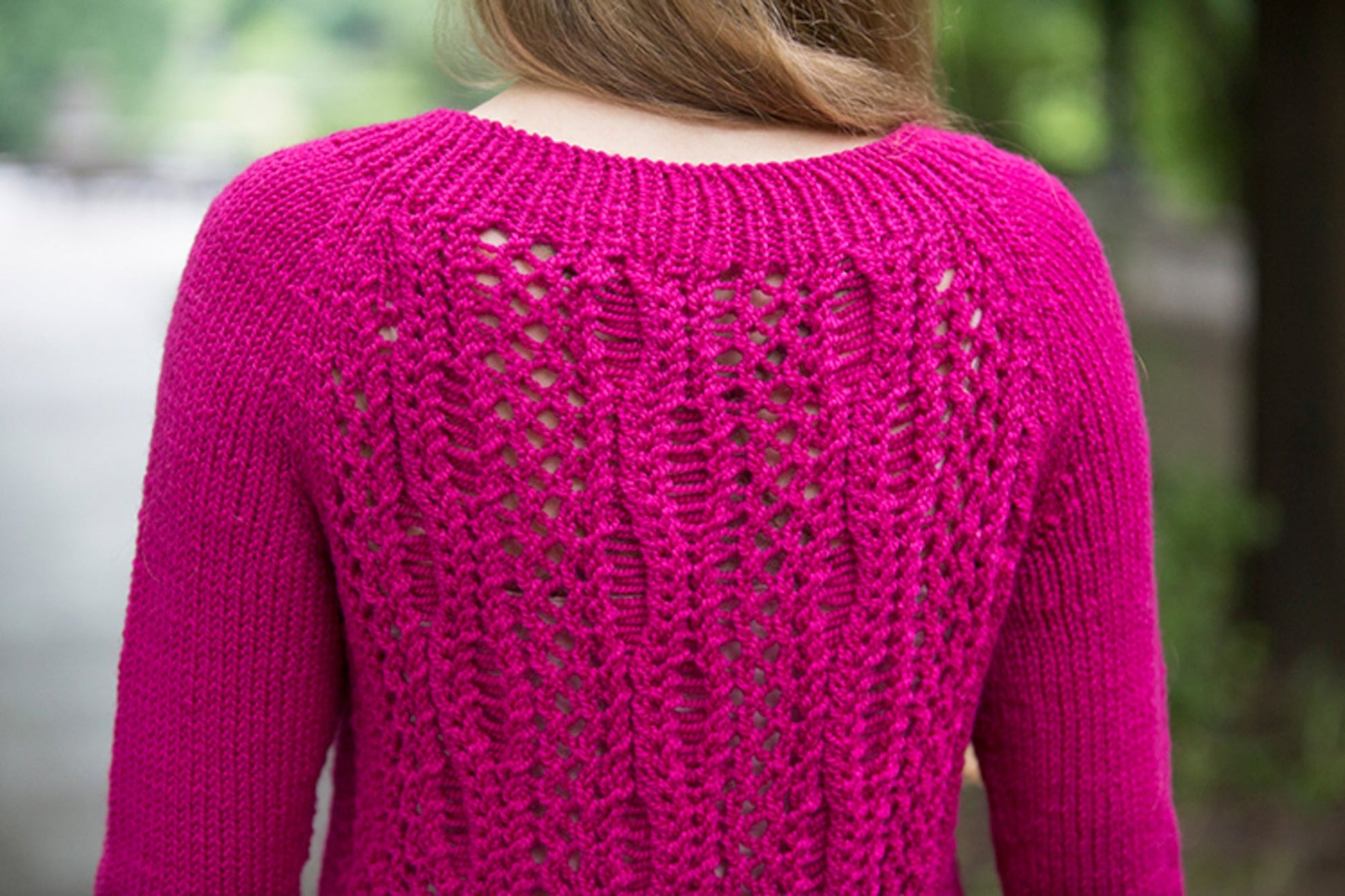 A person with long blond hair is seen from the back wearing a bright pink knitted sweater, much like Halcyon Yarn's Printed Pattern Roxton Pullover. The sweater features an intricate lace pattern on the back, detailed with dropped and twisted stitches, set against a blurred natural green background.