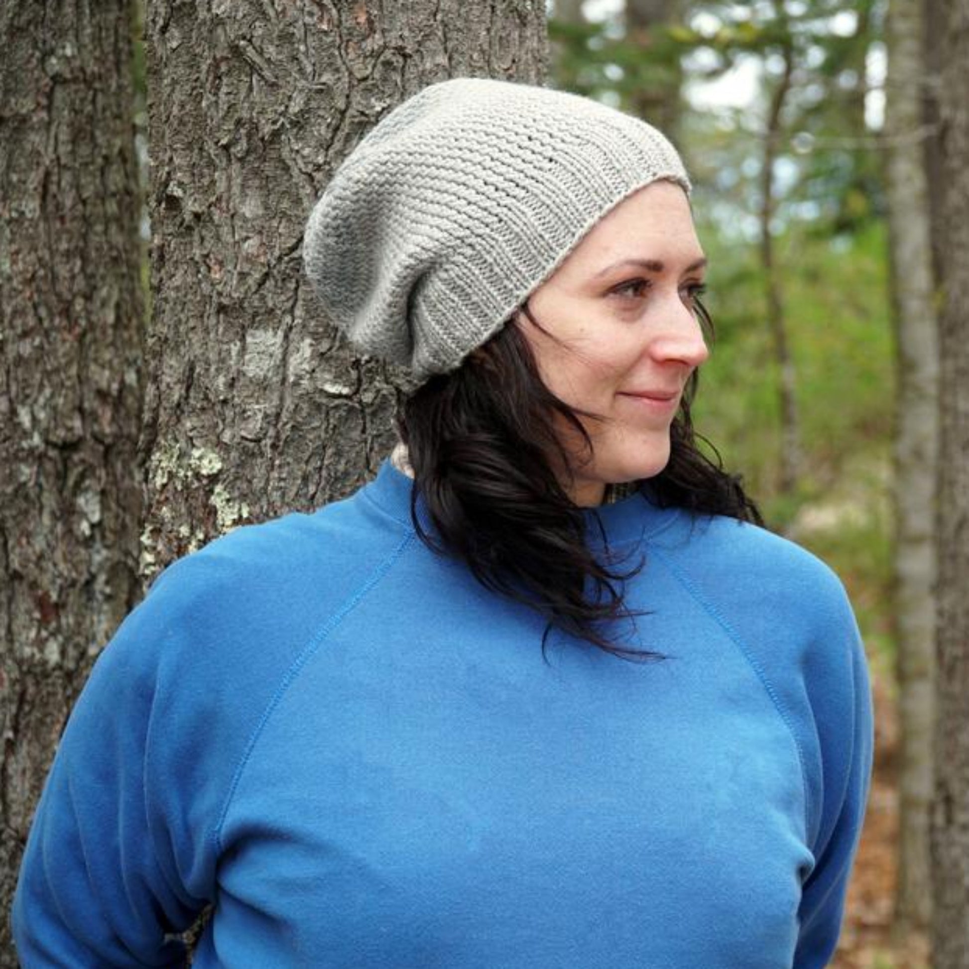 A person wearing a gray Whale Watch Beanie from Halcyon Yarn and a blue sweatshirt stands outdoors, leaning against a tree. The background features additional trees and a natural setting, creating a peaceful and serene atmosphere. The person has dark hair and is smiling gently.