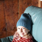 A young child with light hair and pink streaks smiles while sitting on a green couch, wearing a slouchy Whale Watch Beanie from Halcyon Yarn and a red striped sweater. The background is wooden paneling.