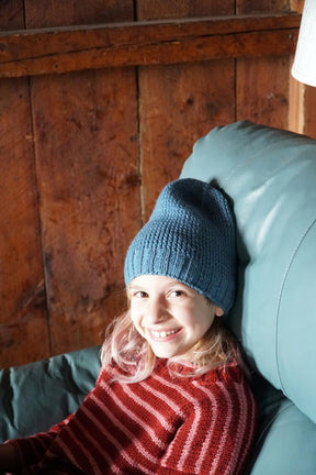 A young child with light hair and pink streaks smiles while sitting on a green couch, wearing a slouchy Whale Watch Beanie from Halcyon Yarn and a red striped sweater. The background is wooden paneling.