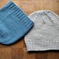 Two Whale Watch Beanies from Halcyon Yarn laid flat on a wooden surface. One slouchy beanie is blue with a ribbed edge, and the other cozy hat is light gray with a similar ribbed edge. The texture of the knit is prominently visible, and both are one size fits all.