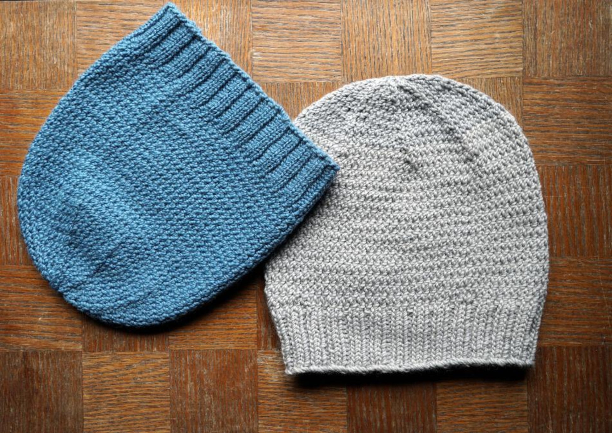 Two Whale Watch Beanies from Halcyon Yarn laid flat on a wooden surface. One slouchy beanie is blue with a ribbed edge, and the other cozy hat is light gray with a similar ribbed edge. The texture of the knit is prominently visible, and both are one size fits all.