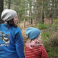 Two people wearing cozy Whale Watch Beanies from Halcyon Yarn walk through a forest. The person on the left sports a blue jacket with an illustration and the word "Maine" on the back, while their look is topped with a slouchy beanie. The individual on the right, with shoulder-length light pink hair, wears a red and white striped sweater.