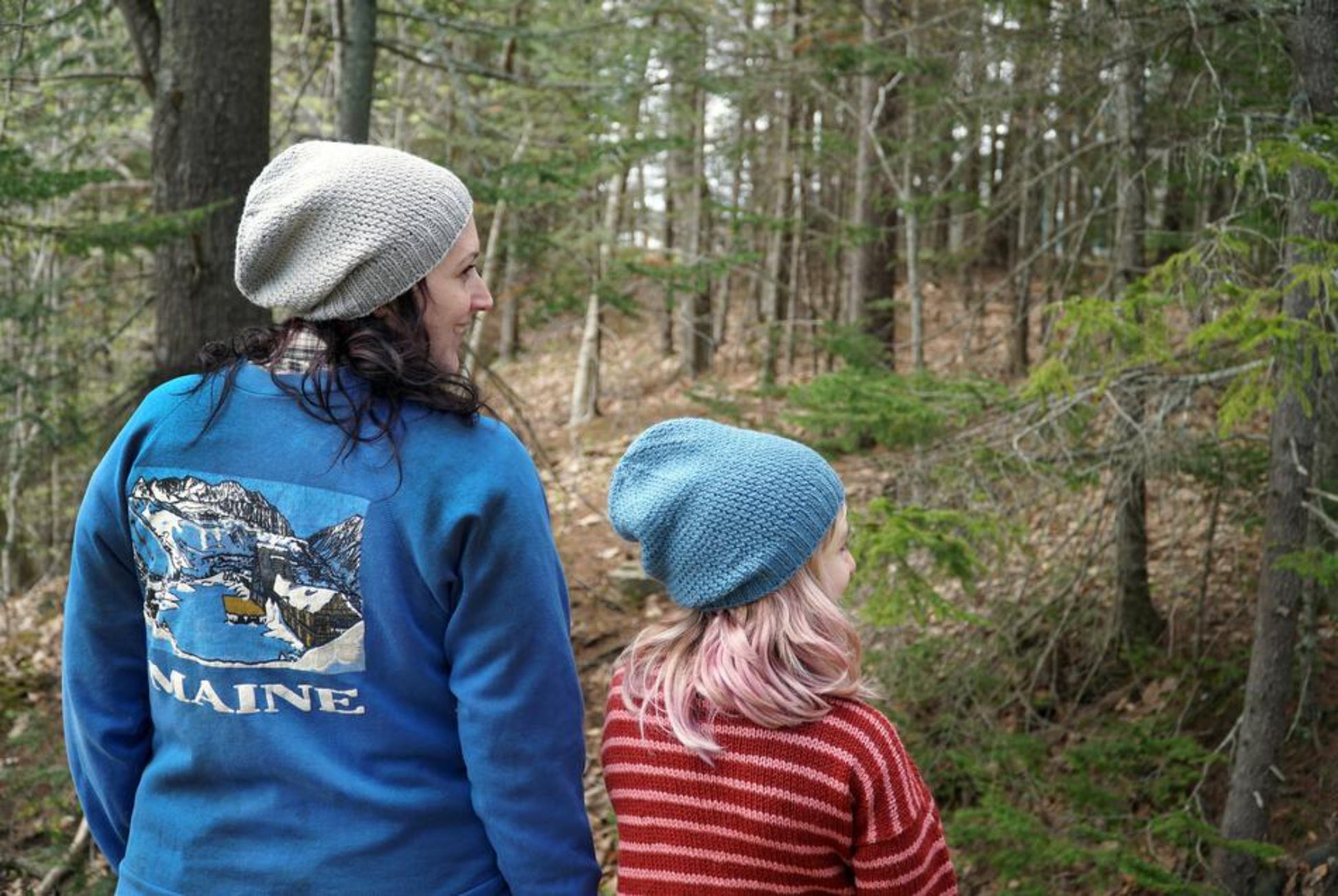 Two people wearing cozy Whale Watch Beanies from Halcyon Yarn walk through a forest. The person on the left sports a blue jacket with an illustration and the word "Maine" on the back, while their look is topped with a slouchy beanie. The individual on the right, with shoulder-length light pink hair, wears a red and white striped sweater.