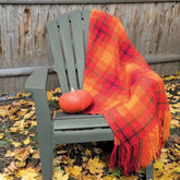 A green Adirondack chair sits outdoors surrounded by fallen yellow leaves. Draped over the chair's back and armrest is the Happy Plaid Woven Blanket from Halcyon Yarn, featuring a striking red and orange pattern. On the seat rests a small orange pumpkin, while a wooden fence provides the backdrop.