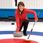 A person wearing a Twist Collective Bonspiel Cardigan Sweater kneels on an ice rink, preparing to launch a curling stone with their right hand and holding a curling broom with their left. They are smiling, and the curling rink features blue, white, and red concentric circles.