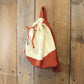 The Halcyon Yarn Auditorium Knitted Backpack in beige and rust colors hangs on a rustic, wooden wall. The bag is predominantly beige with a rust-colored bottom and matching drawstring closure tied at the top. The wood wall has a textured, natural appearance with visible nails and wood grain.