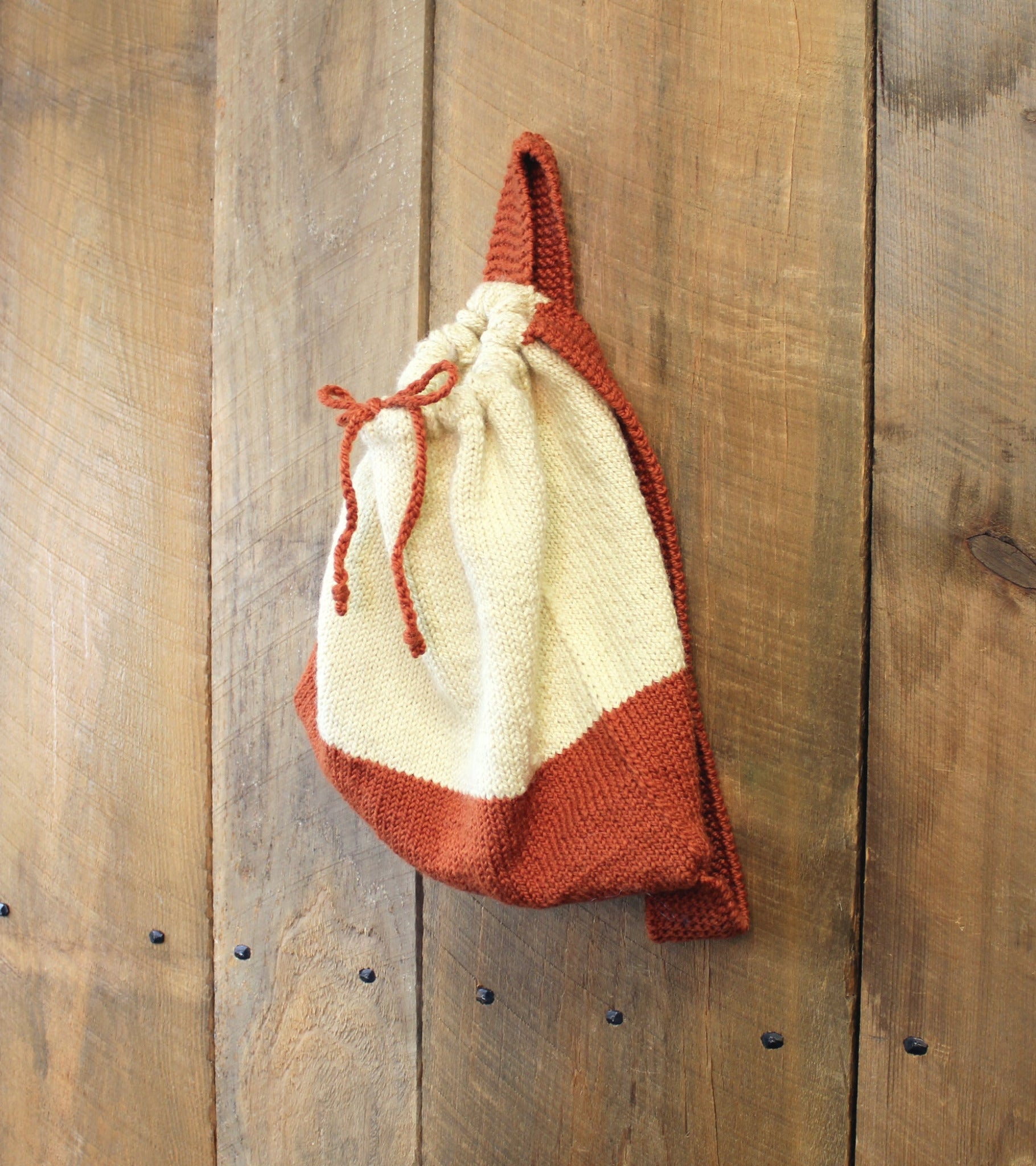 The Halcyon Yarn Auditorium Knitted Backpack in beige and rust colors hangs on a rustic, wooden wall. The bag is predominantly beige with a rust-colored bottom and matching drawstring closure tied at the top. The wood wall has a textured, natural appearance with visible nails and wood grain.