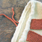 A close-up of the Auditorium Knitted Backpack by Halcyon Yarn on a wooden surface. The backpack features cream and burnt orange yarn with a drawstring closure made of burnt orange yarn. The texture and stitching of the knitted fabric are clearly visible, showcasing intricate craftsmanship reminiscent of Norumbega traditions.