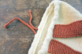 A close-up of the Auditorium Knitted Backpack by Halcyon Yarn on a wooden surface. The backpack features cream and burnt orange yarn with a drawstring closure made of burnt orange yarn. The texture and stitching of the knitted fabric are clearly visible, showcasing intricate craftsmanship reminiscent of Norumbega traditions.