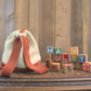 An Auditorium Knitted Backpack from Halcyon Yarn, featuring a drawstring closure and striking orange straps, is placed beside vintage wooden alphabet blocks spelling "HALCYON." This charming scene unfolds on an old wooden tabletop against a backdrop of vertically aligned wooden planks.