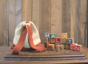 An Auditorium Knitted Backpack from Halcyon Yarn, featuring a drawstring closure and striking orange straps, is placed beside vintage wooden alphabet blocks spelling "HALCYON." This charming scene unfolds on an old wooden tabletop against a backdrop of vertically aligned wooden planks.