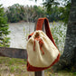 The Auditorium Knitted Backpack by Halcyon Yarn, featuring a red and cream color scheme, hangs on a wooden fence by the serene Norumbega river. With its drawstring closure and long strap for carrying, the background reveals peaceful riverside scenery with trees and subtle greenery.