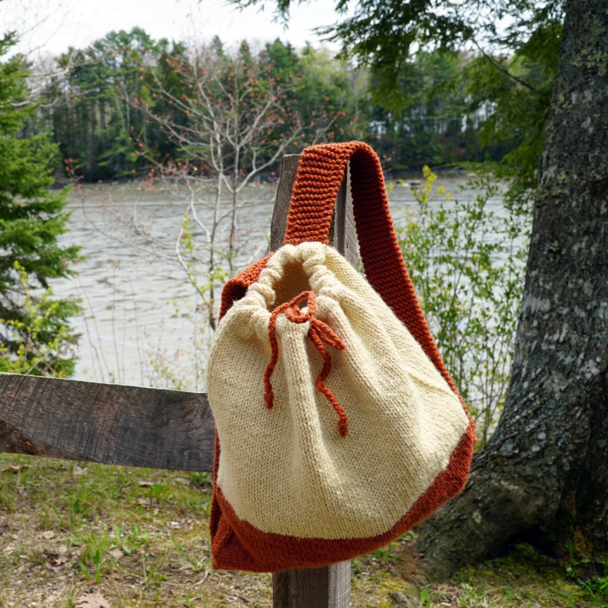 An Auditorium Knitted Backpack by Halcyon Yarn, handcrafted with a drawstring closure from cream and rust-colored yarn, hangs on a wooden fence. Beyond the fence, a river flows surrounded by lush green trees, creating a serene outdoor setting ideal for your next Norumbega adventure.