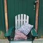 A green wooden Adirondack chair sits on a porch in front of a dark green door. Resting on the chair are two cozy Halcyon Yarn Eastport Pillows, one featuring blue and white woven fabric, the other orange and white. Above the door is a small round brass or gold-colored decorative ring.