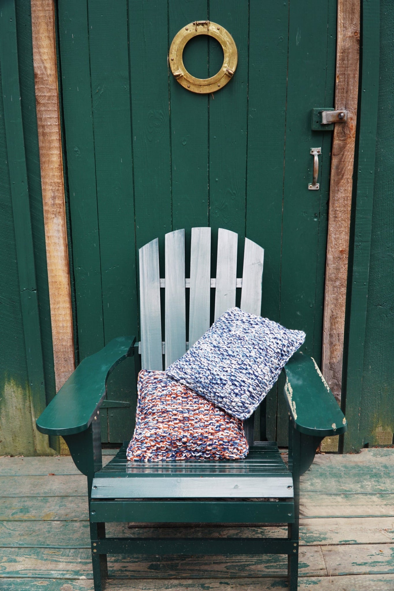 A green wooden Adirondack chair sits on a porch in front of a dark green door. Resting on the chair are two cozy Halcyon Yarn Eastport Pillows, one featuring blue and white woven fabric, the other orange and white. Above the door is a small round brass or gold-colored decorative ring.