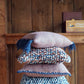 A stack of four Halcyon Yarn's Eastport Pillows, featuring an array of woven designs and colors akin to a knitting project, in blue and earthy tones, is arranged on a green wooden stool against a rustic wooden wall. In the background, a small shelf holds a box labeled 'Lighthouse Crackers' and a dried bunch of herbs.